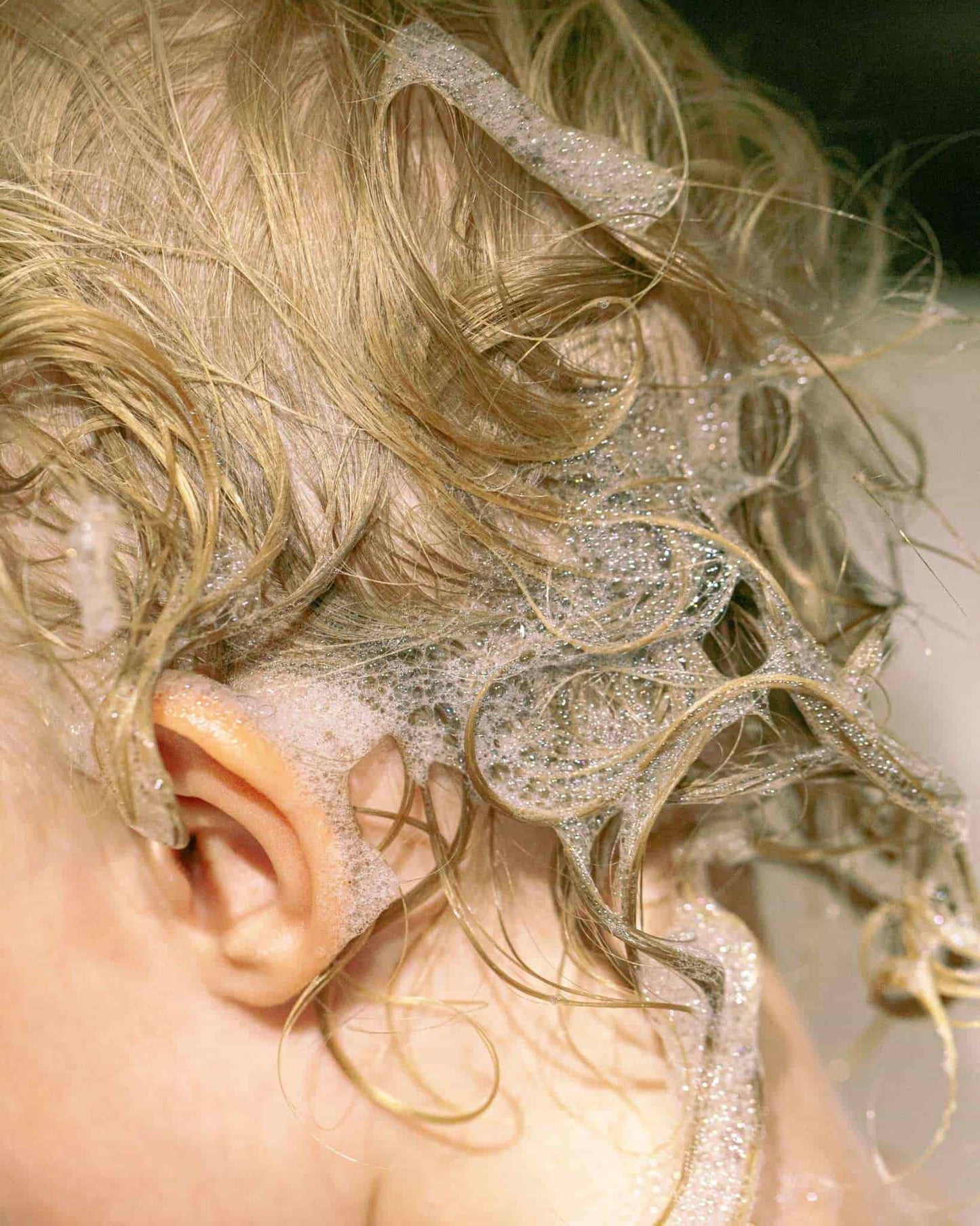 Close-up of a child's head with wet, blonde hair and soapy suds on a neutral background