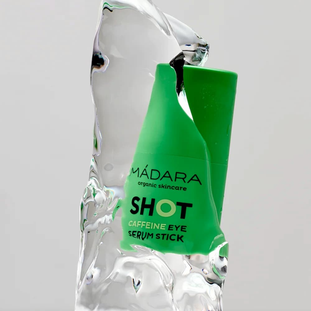 A green MáDARA skincare serum stick labeled 'SHOT Caffeine Eye Serum Stick' emerging from clear, sculpted ice against a light gray background.